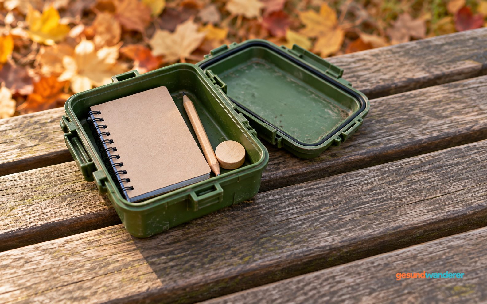 Geöffnete Geocache-Dose mit Notizbuch, Bleistift und Holzplättchen auf einer Bank